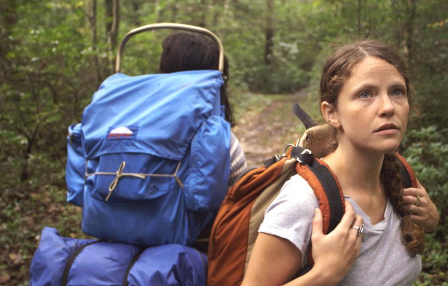Woman stands in a forest holding the straps of her orange backpack.
